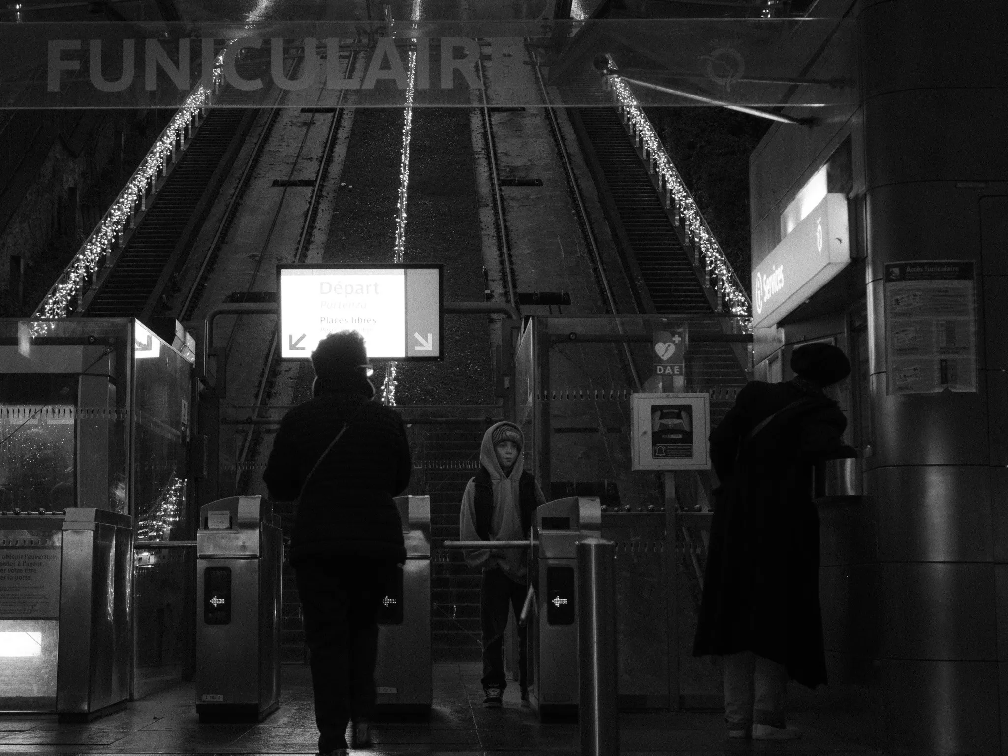 Black and white photo of people passing the ticket gates at a funicular station, the tracks rising behind them lined with string lights.