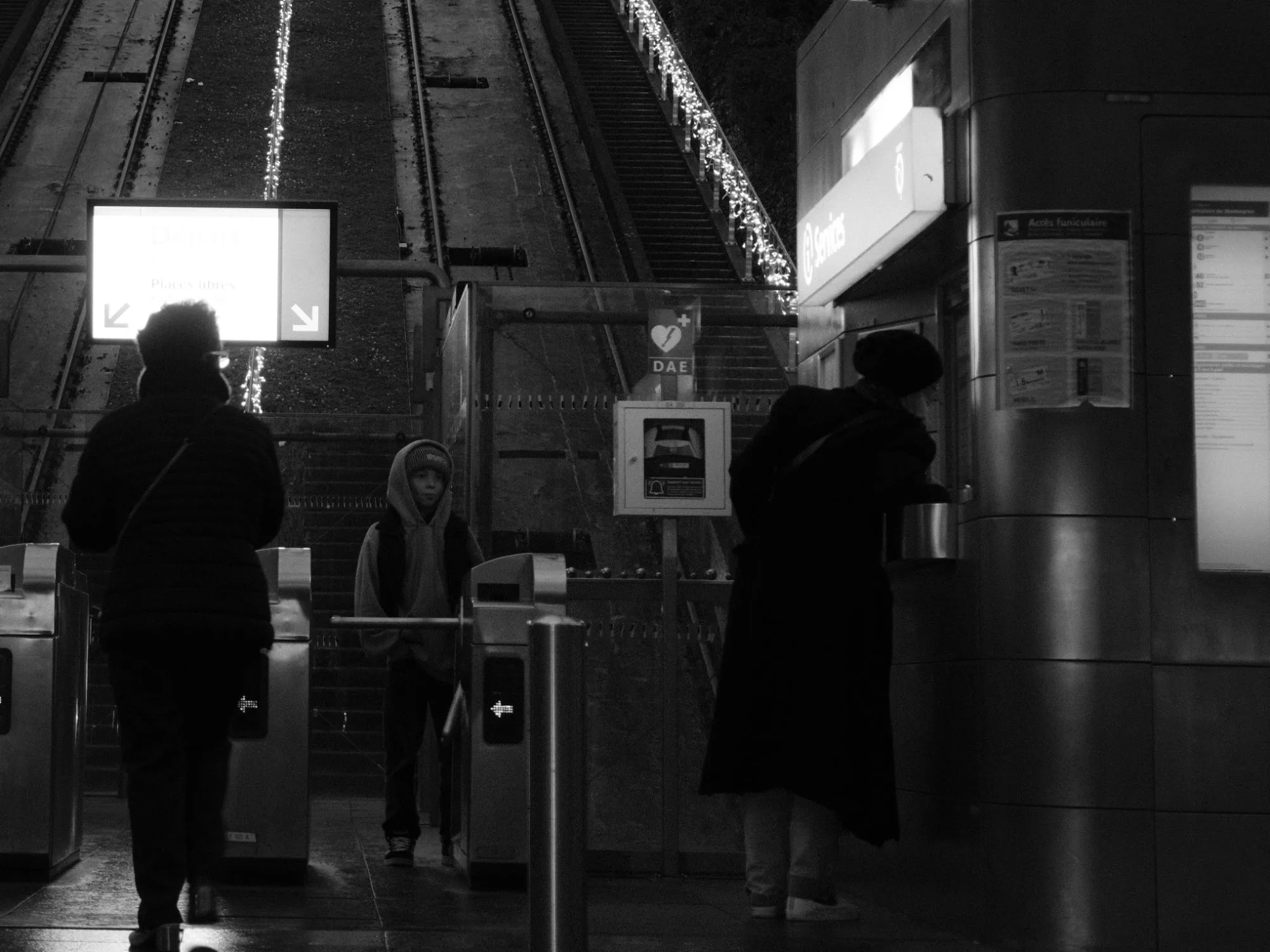 Black and white photo of people passing the ticket gates at a funicular station, the tracks rising behind them lined with string lights.