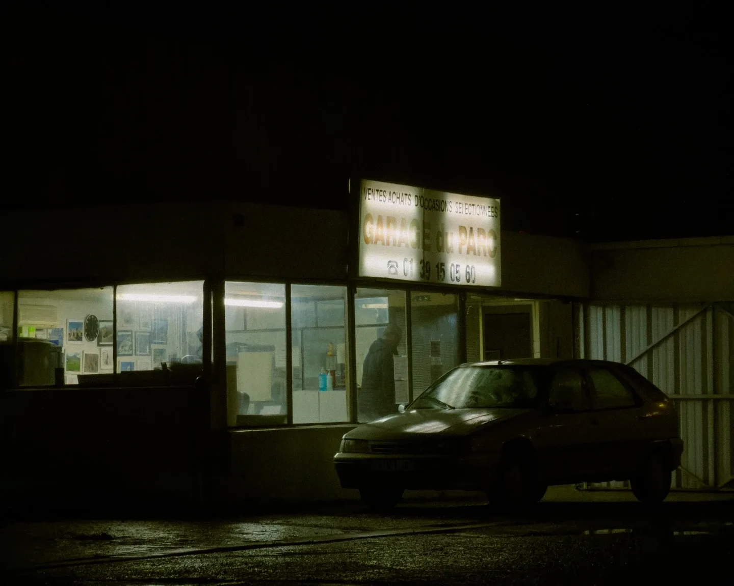 A lit-up used car garage at night, its “Garage du Parc” sign glowing above a parked hatchback.