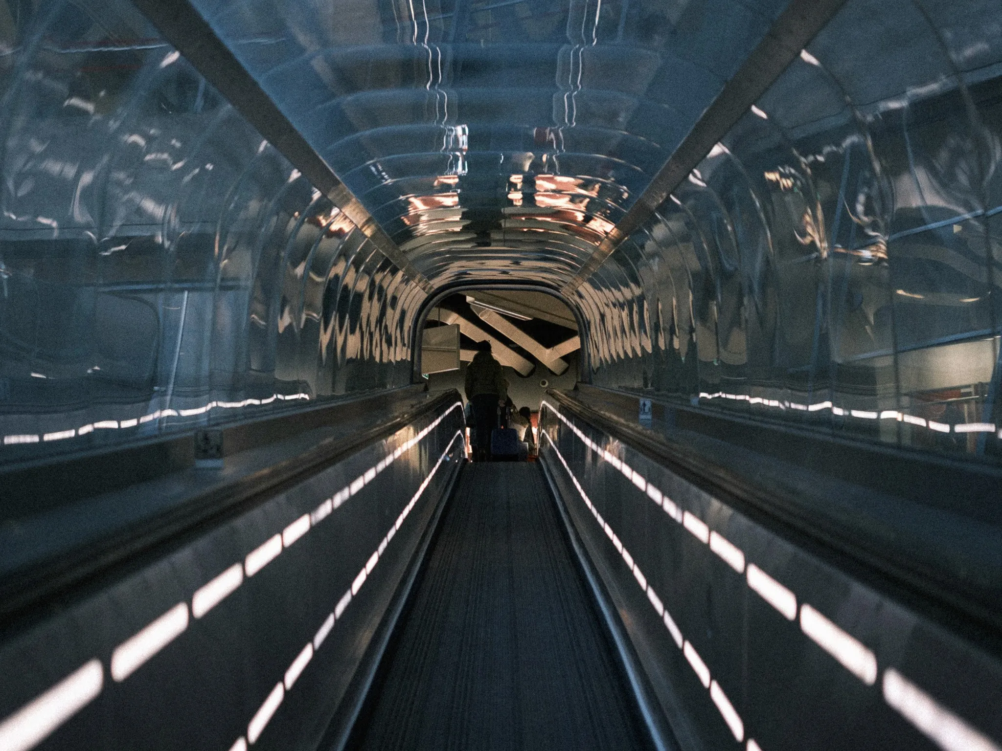 A moving walkway running through a reflective glass tube towards a lone traveller in the distance.