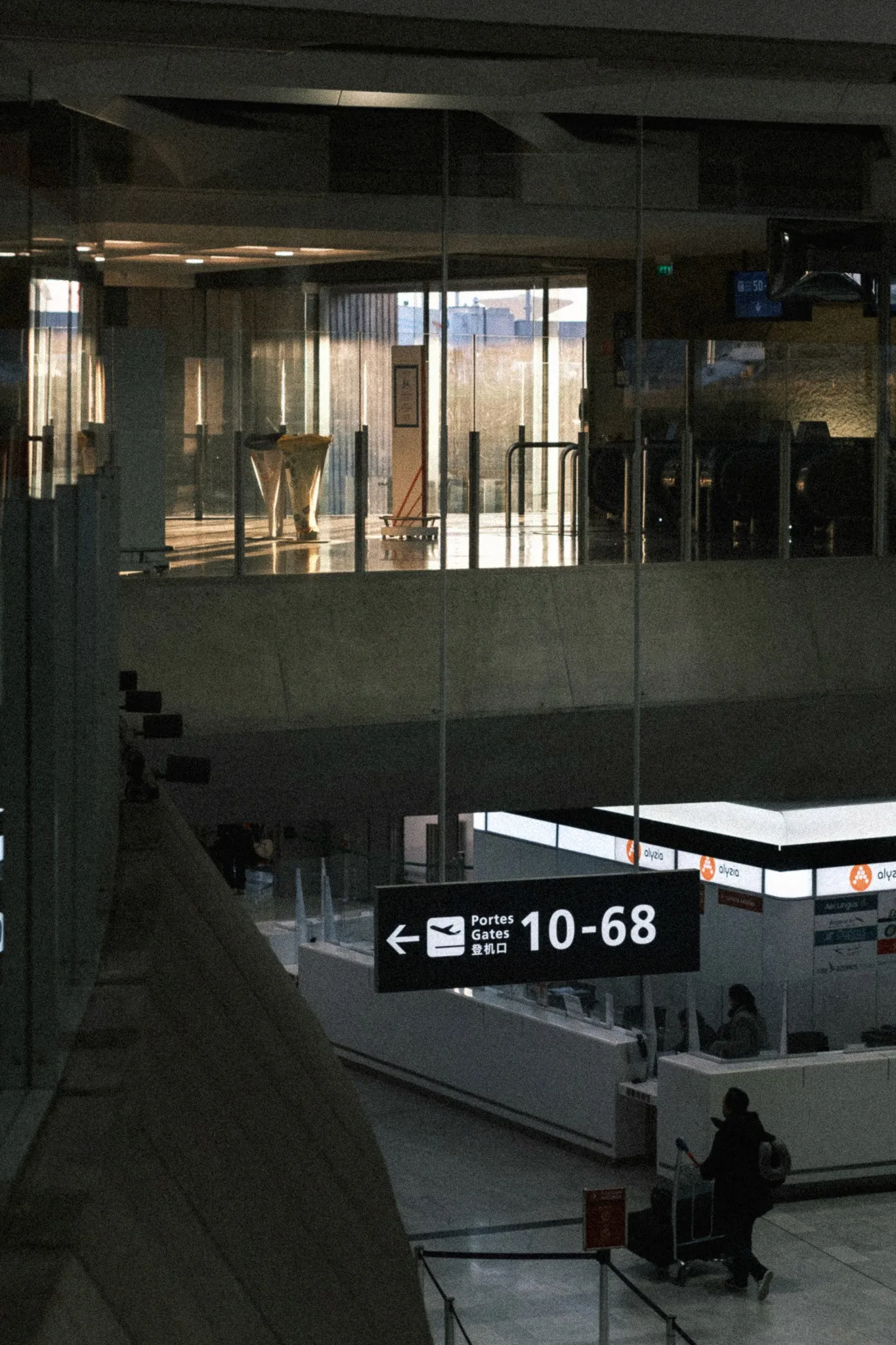 Looking down into an airport terminal at dusk, a sign pointing to gates 10–68 above a traveller pushing a luggage trolley.
