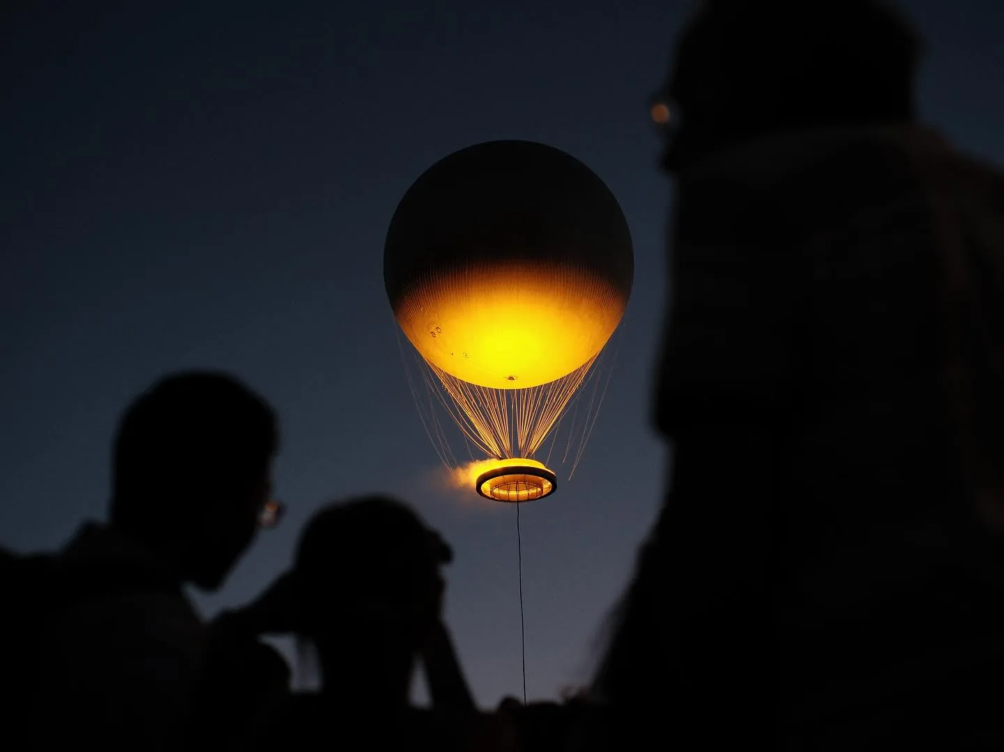 A glowing golden balloon rising into the dusk sky above the silhouettes of onlookers.