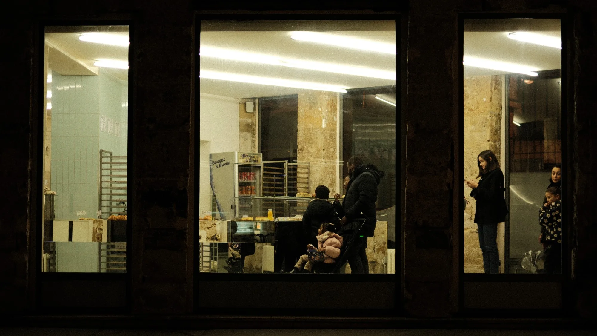 Three bakery windows at night framing customers waiting under fluorescent light.