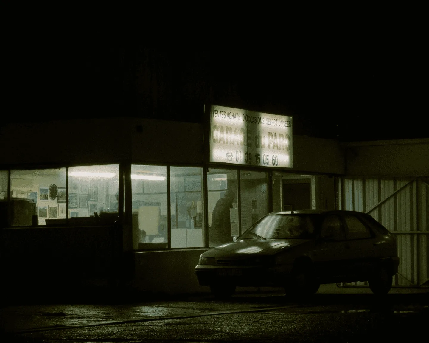 A lit-up used car garage at night, its “Garage du Parc” sign glowing above a parked hatchback.