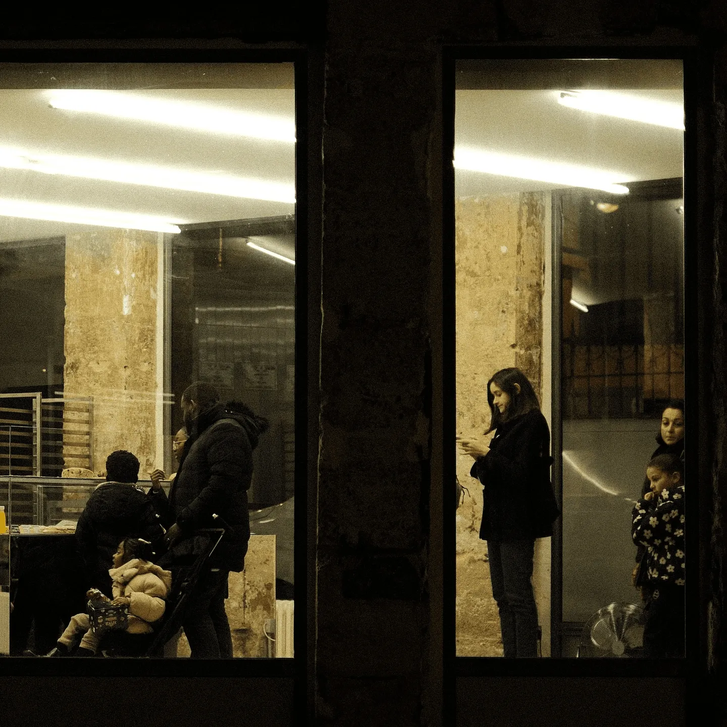 Two bakery windows at night framing customers waiting under fluorescent light.