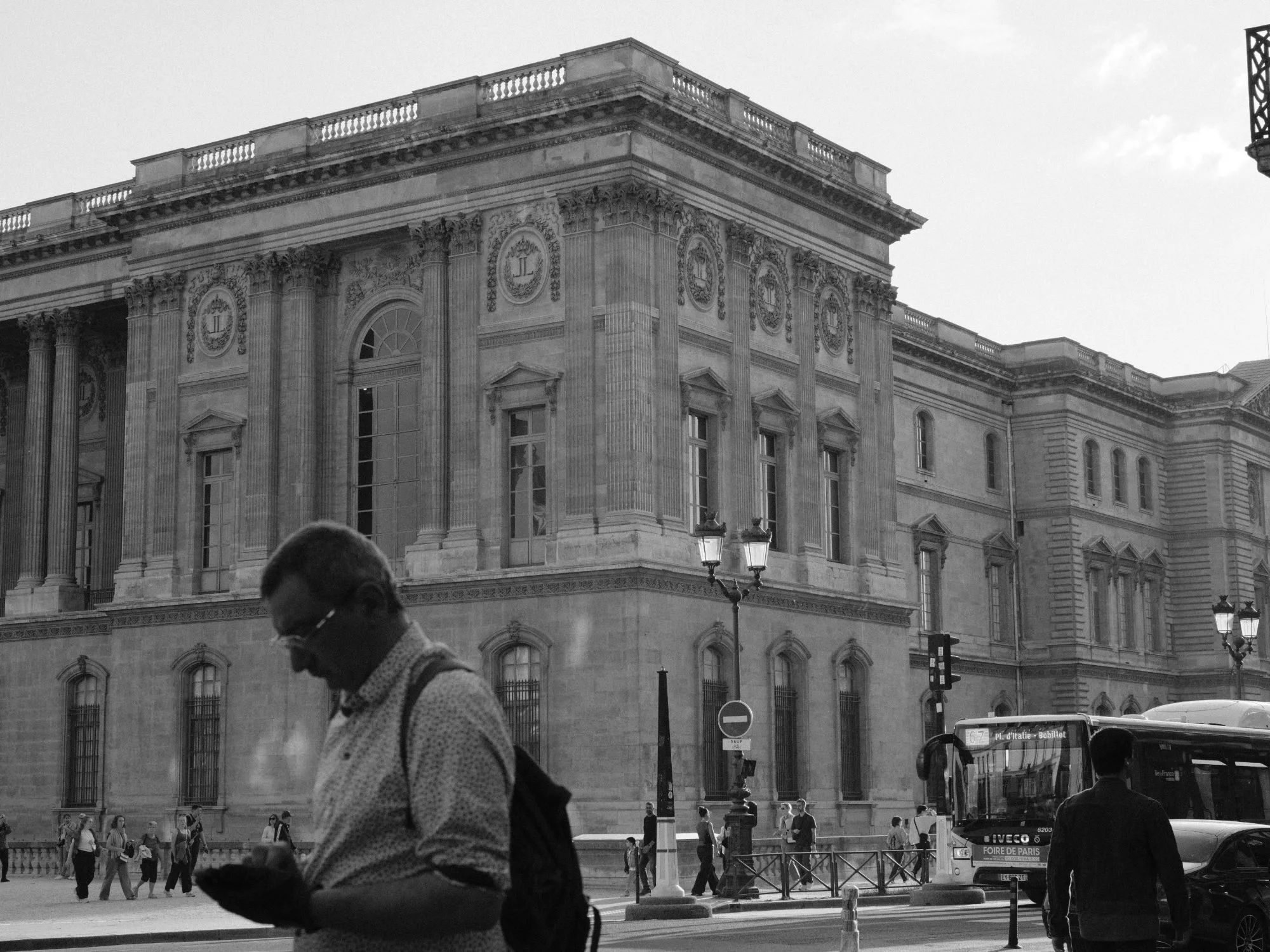 Black and white street scene of a man looking at his phone in front of the ornate façade of the Louvre.