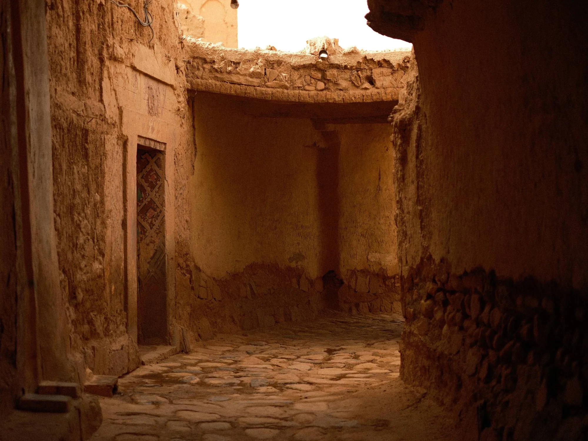 A narrow cobbled alley winding between mud-brick walls, a patterned wooden door set into one side.