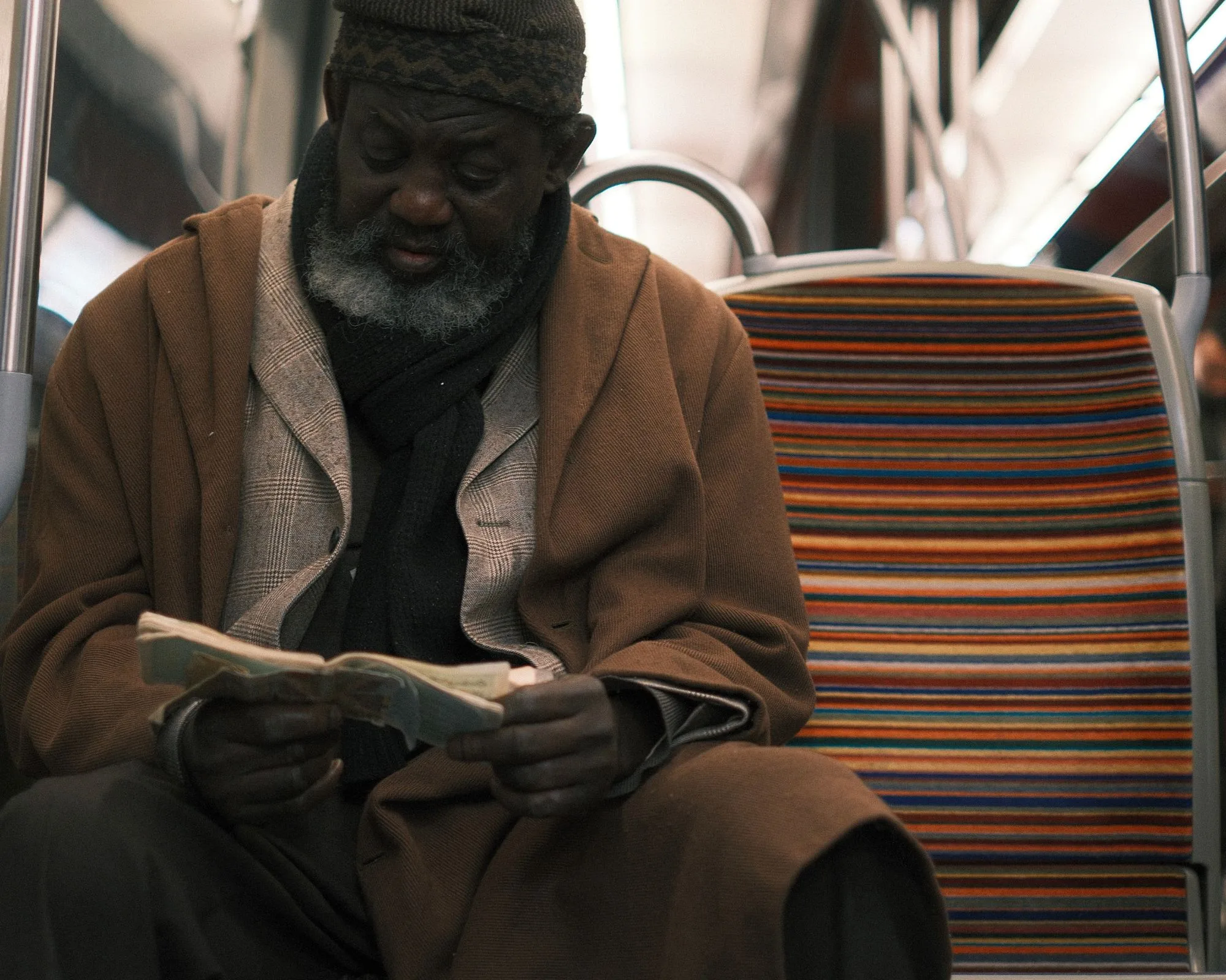 An older man in a brown coat and knitted cap reading a worn booklet on the metro, beside an empty seat with bright striped upholstery.